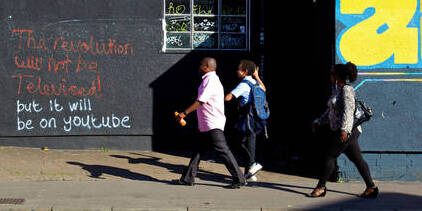两名成年人和一名学童走在街上，路过涂鸦，上面写着“革命不会被电视转播，但会在YouTube上。”